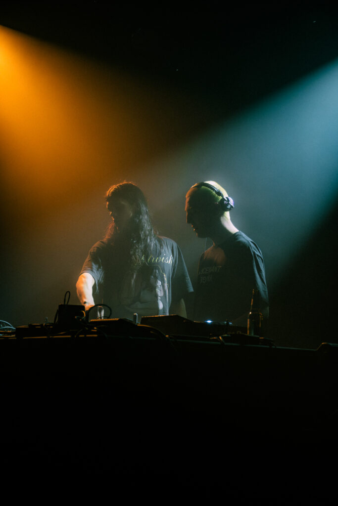 Zwei DJs bei einem Live-Event mit dramatischer Bühnenbeleuchtung und dunkler Konzertatmosphäre.
