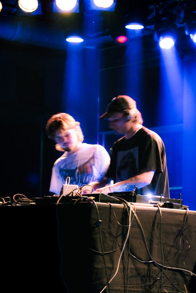 Zwei DJs performen live am Mischpult unter blauen Bühnenlichtern bei einem Musik-Event.