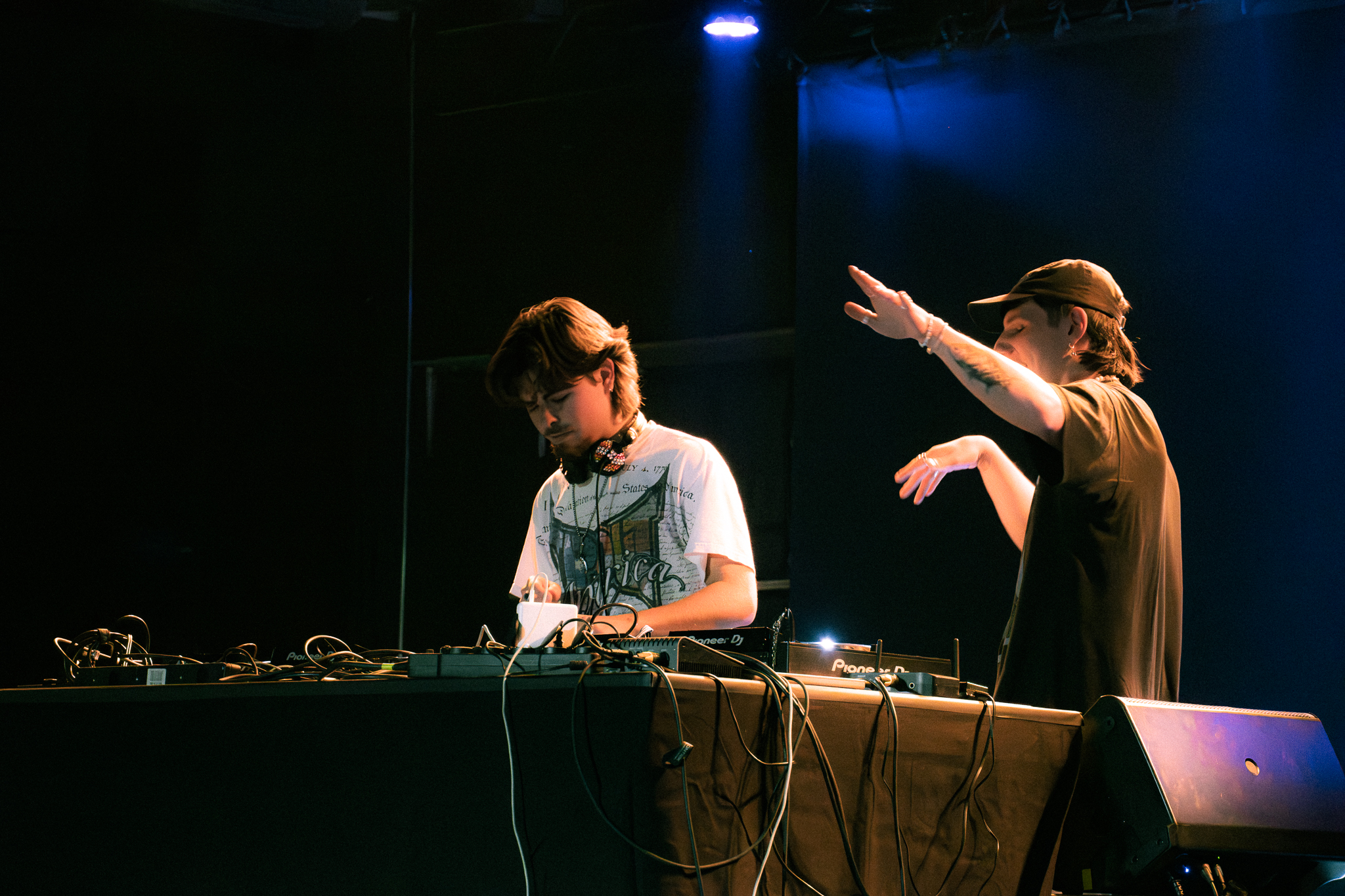 Zwei DJs performen live auf einer Bühne mit blauem Licht und DJ-Equipment während eines Club-Events.