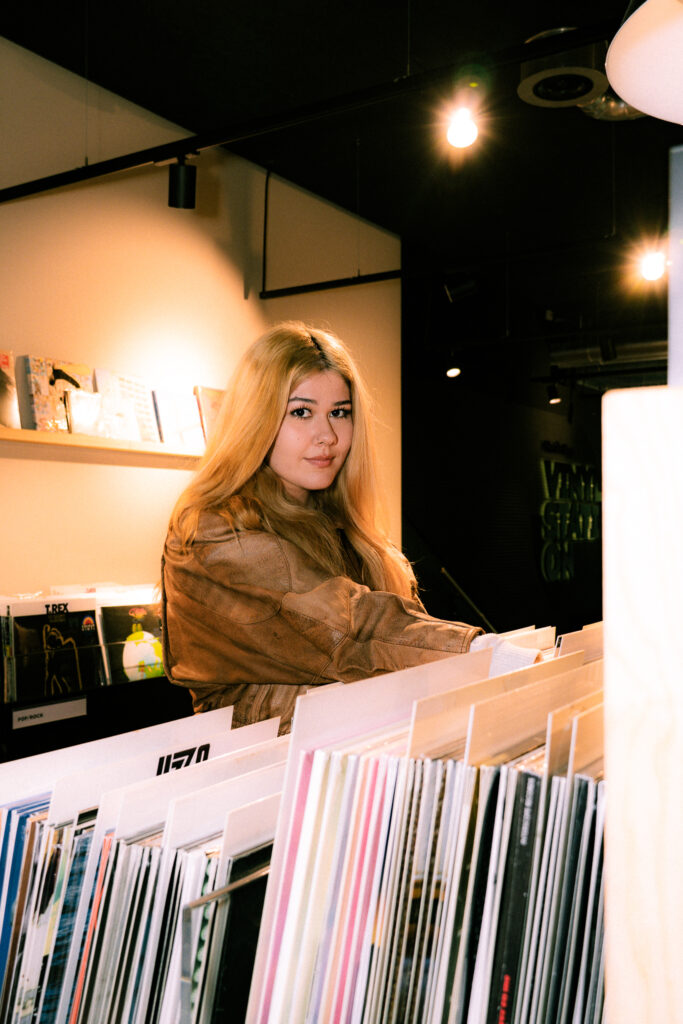 Eine blonde Frau schaut lächelnd über Schallplatten hinweg in die Kamera, während warmes Licht den Plattenladen erhellt.